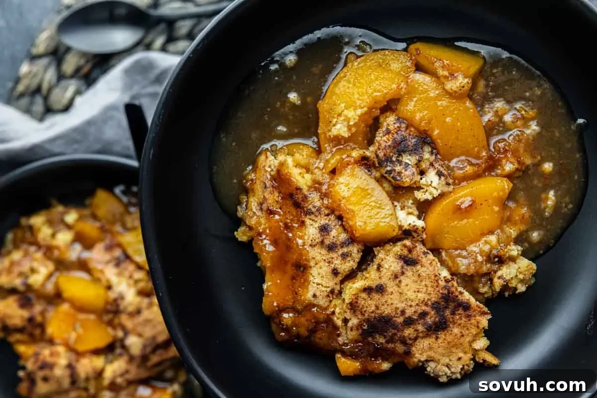 Two black bowls filled with peach cobbler, featuring baked peaches and golden brown crust, are placed on a dark surface with a spoon nearby.