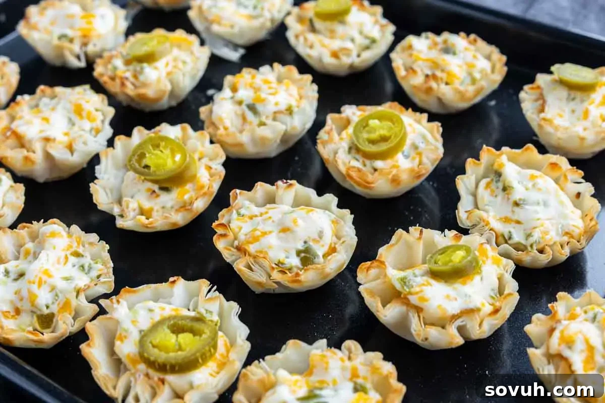 Mini phyllo pastry cups filled with melted cheese and topped with sliced jalapeños, arranged on a black baking tray.