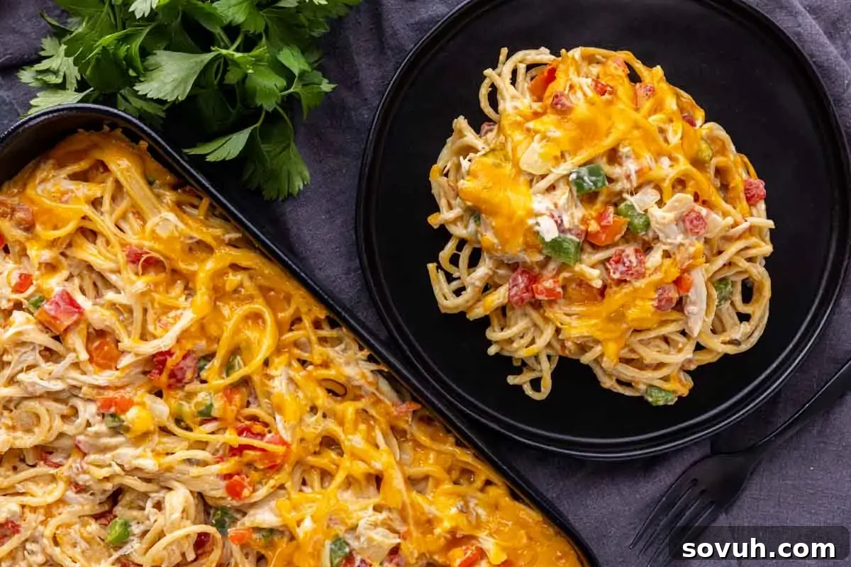 A plate and baking dish of cheesy chicken spaghetti with visible vegetables, topped with melted cheddar cheese, next to fresh parsley.