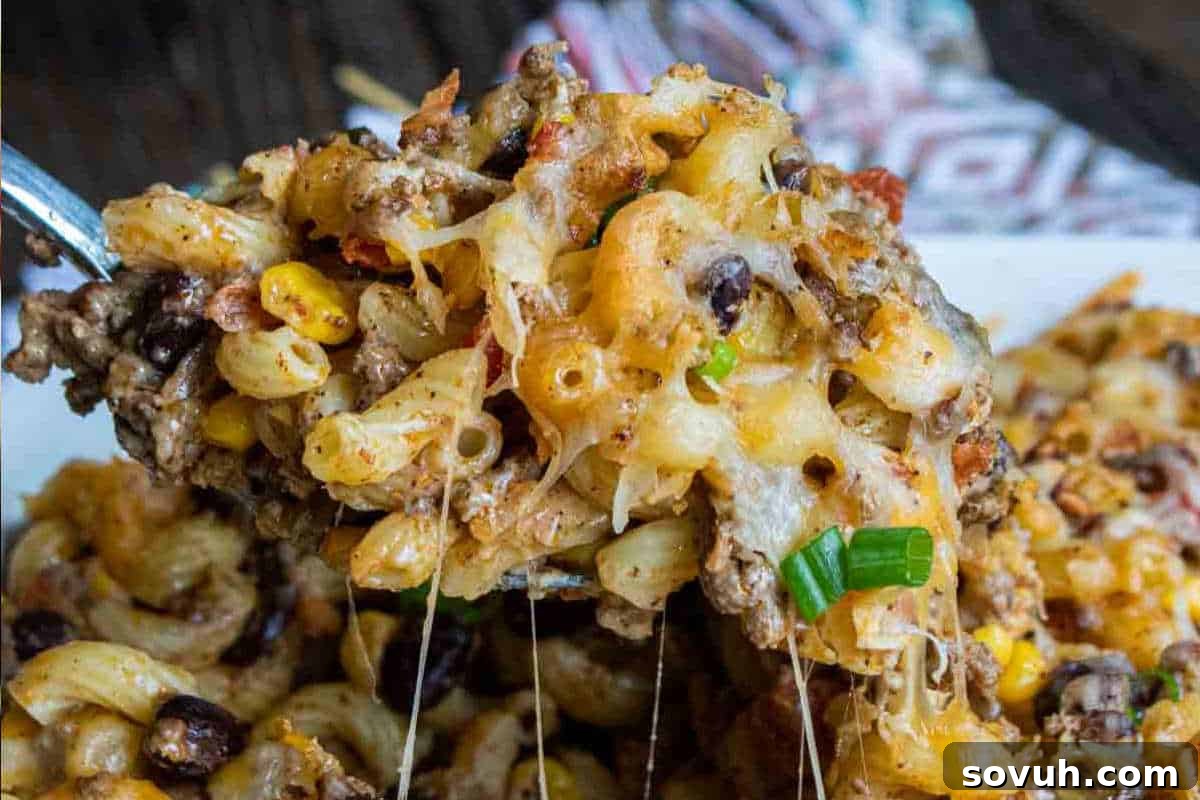 A close-up of a cheesy pasta bake with ground beef, black beans, corn, green onions, and melted cheese being scooped with a spoon.