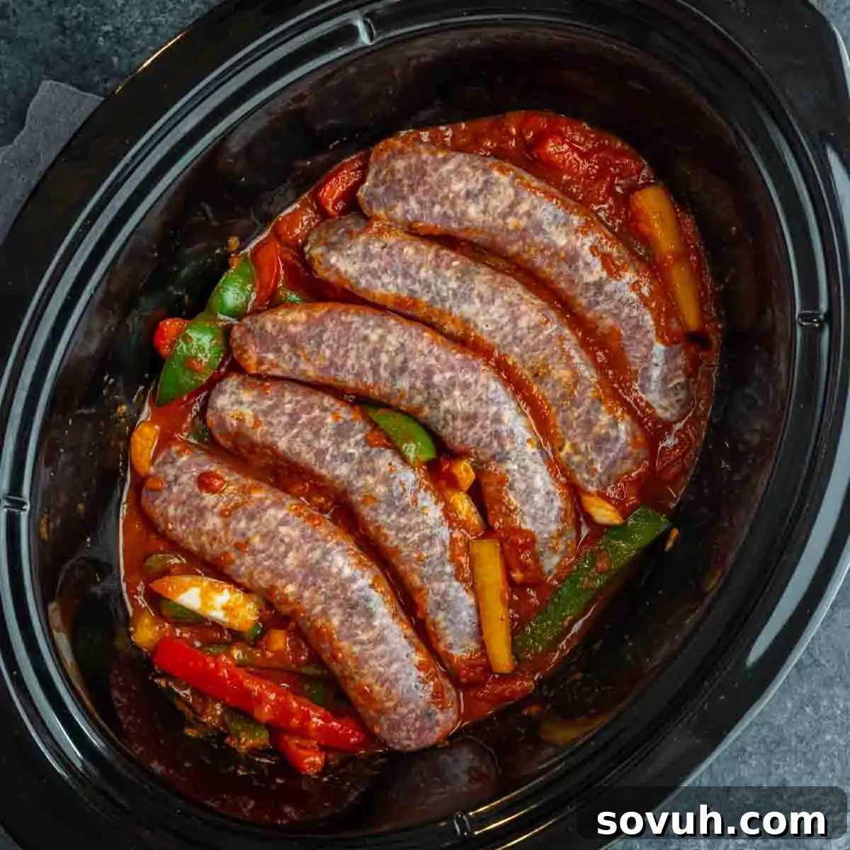 Five raw sausages are placed on top of tomato sauce with sliced bell peppers in a slow cooker, making the perfect base for a flavorful Italian sausage and peppers slow cooker recipe.
