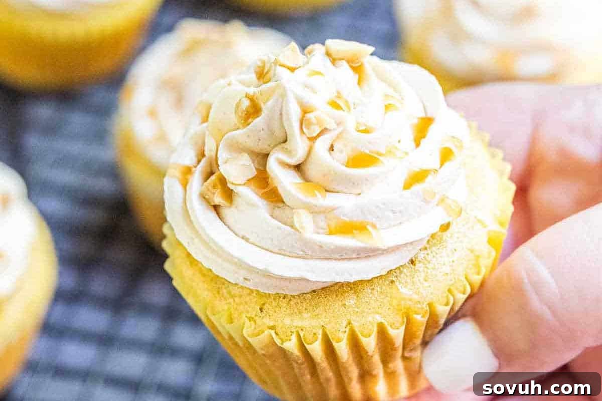 A hand gently holds a vanilla cupcake, exquisitely frosted with a swirled buttercream, adorned with a glossy caramel drizzle and a sprinkle of crushed nuts. More inviting cupcakes are softly blurred in the background.