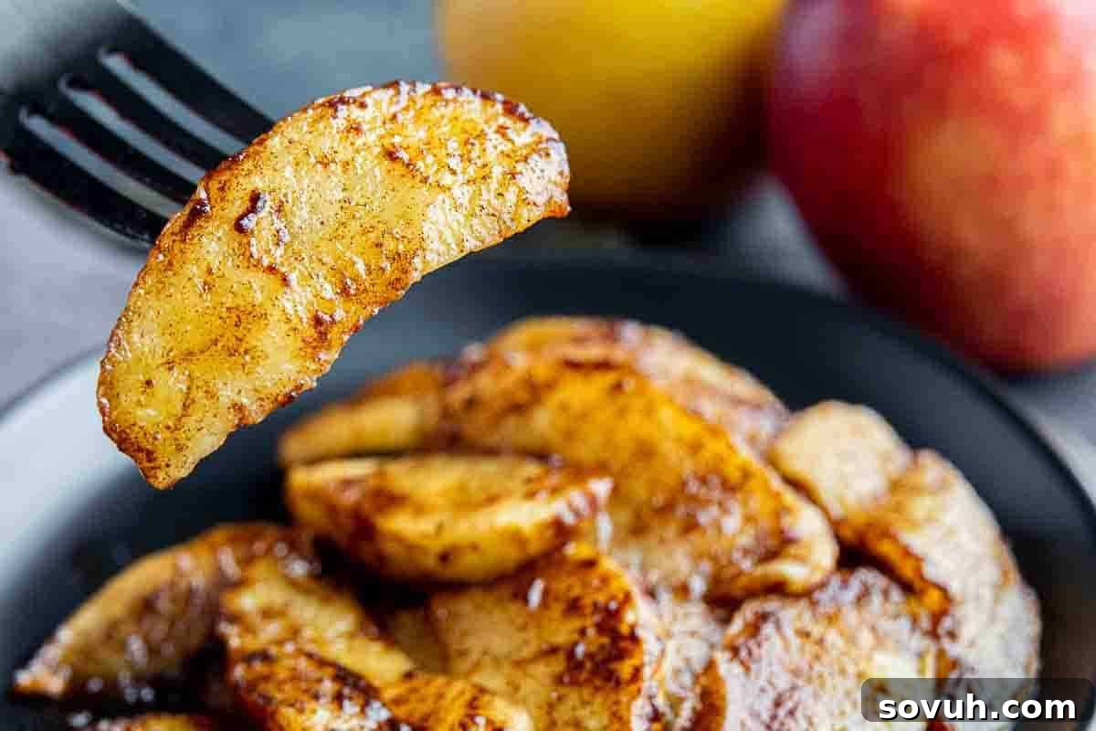 A close-up view of a fork holding a perfectly cooked cinnamon apple slice, lifted above a plate adorned with more glistening apple slices. Whole apples, softly blurred in the background, underscore the fresh, cozy appeal of this fall recipe.
