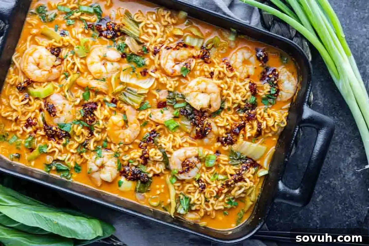 A rectangular black pan filled with a spicy shrimp ramen bake, garnished with herbs, sits on a dark countertop beside fresh green onions and leafy greens.