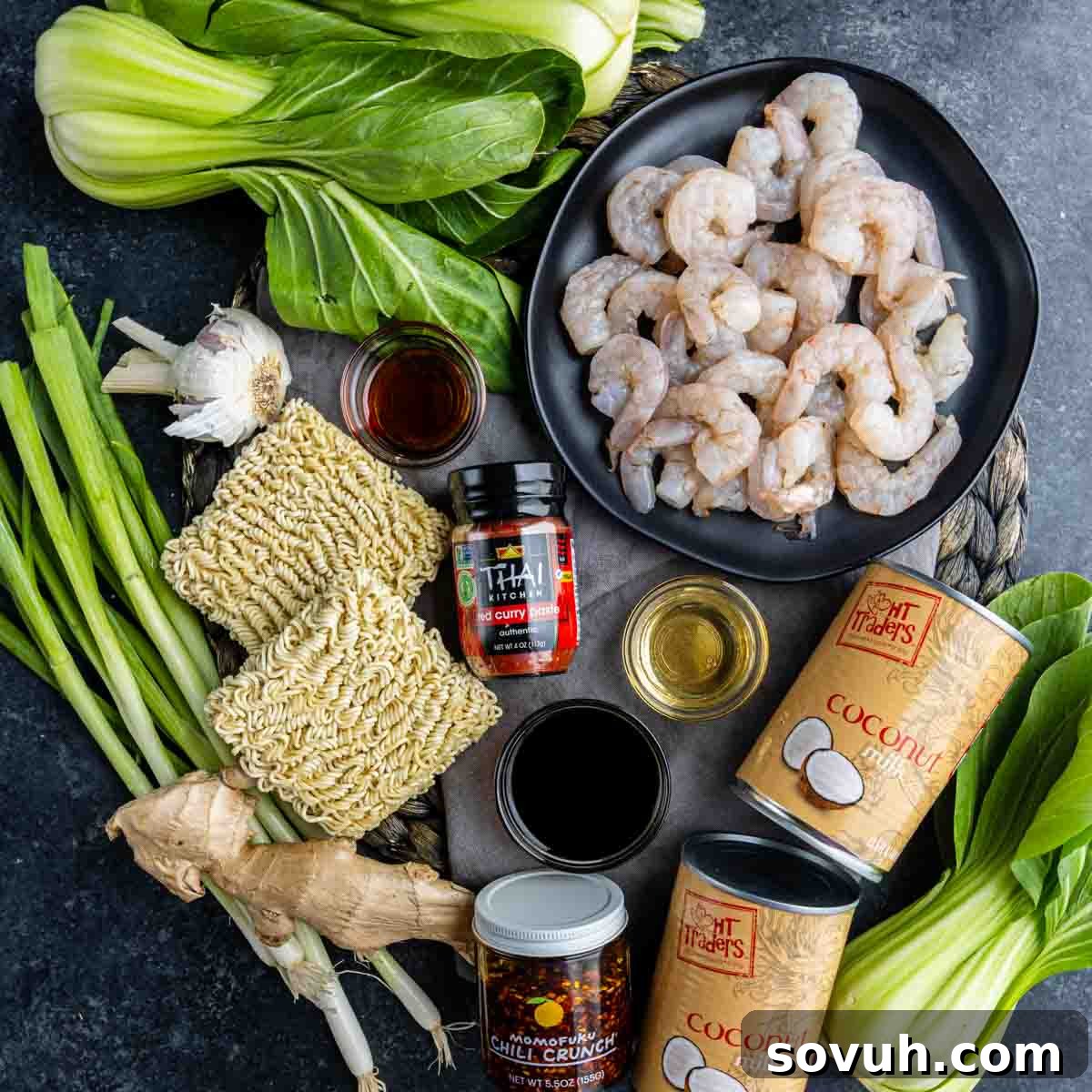 Ingredients for a Shrimp Ramen Bake are arranged on a surface, including shrimp, bok choy, ramen noodles, garlic, sauces, ginger, coconut milk, and seasonings.