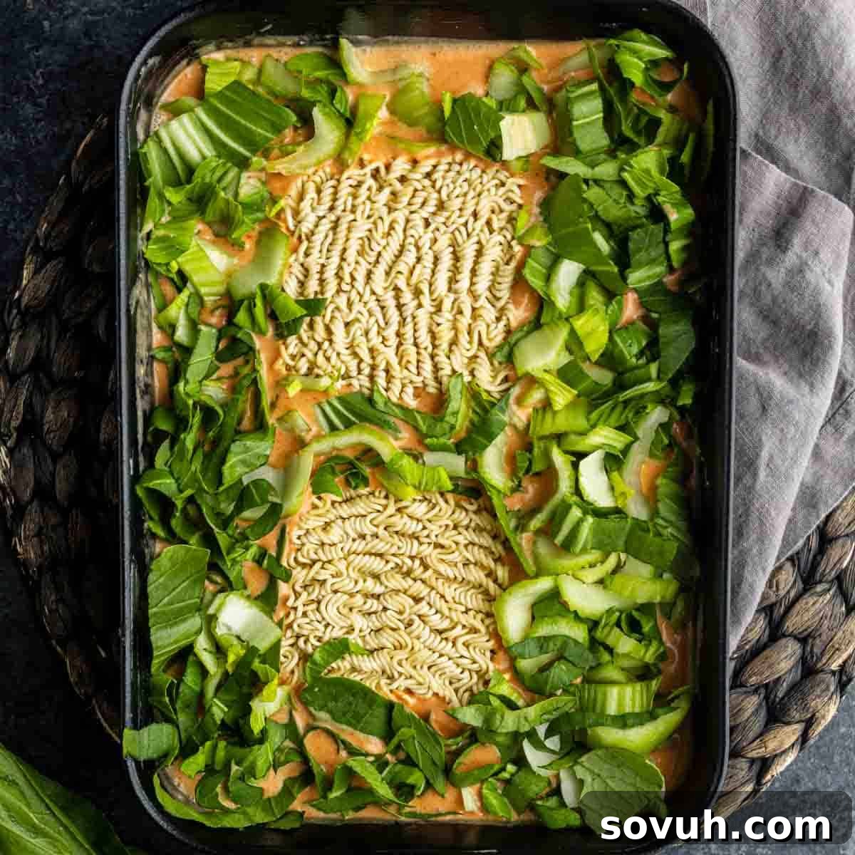 Uncooked instant ramen noodles in a baking dish with chopped bok choy and celery, surrounded by a creamy orange sauce—an easy ramen casserole perfect for busy weeknights.