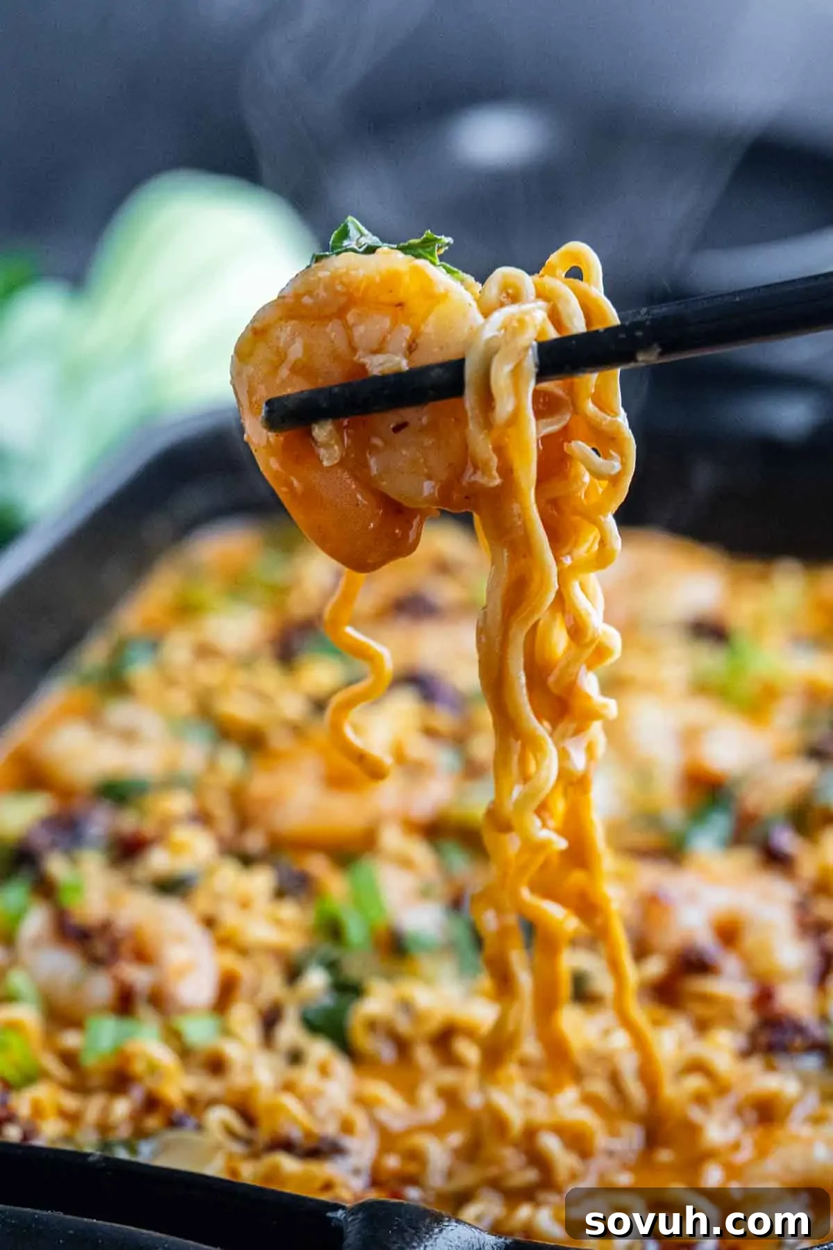 Chopsticks holding shrimp and ramen noodles above a bowl of soup, reminiscent of a Shrimp Ramen Bake, with visible green onions and broth in the background.