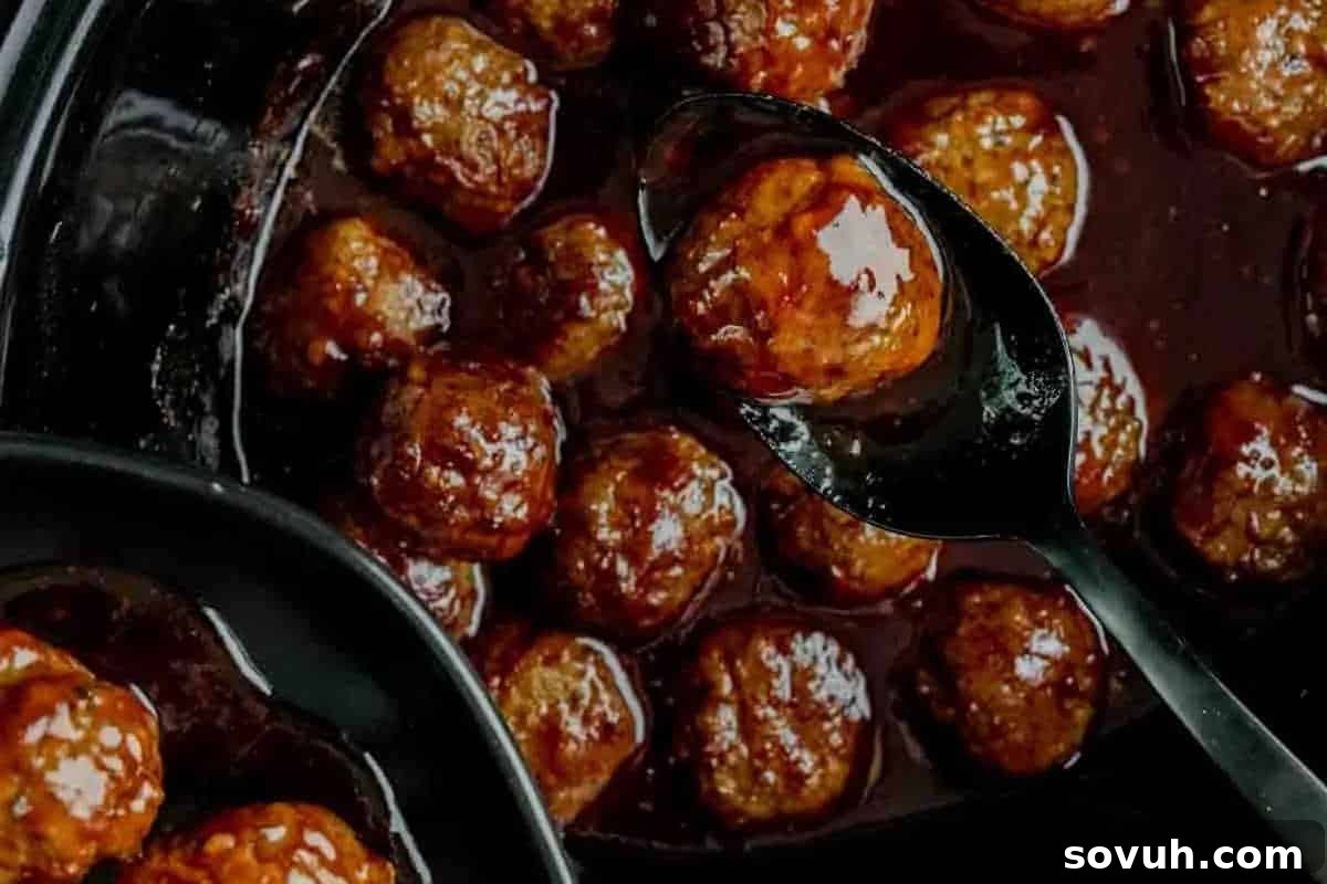 A spoon lifting a perfectly glazed Crock Pot Meatball from a dish filled with several other savory-sweet meatballs in a dark sauce.