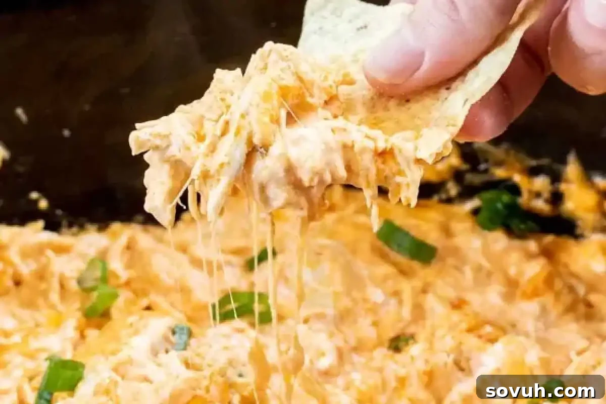 Close-up of a hand holding a tortilla chip dipped into creamy, cheesy Crock Pot Buffalo Chicken Dip, garnished with green herbs.