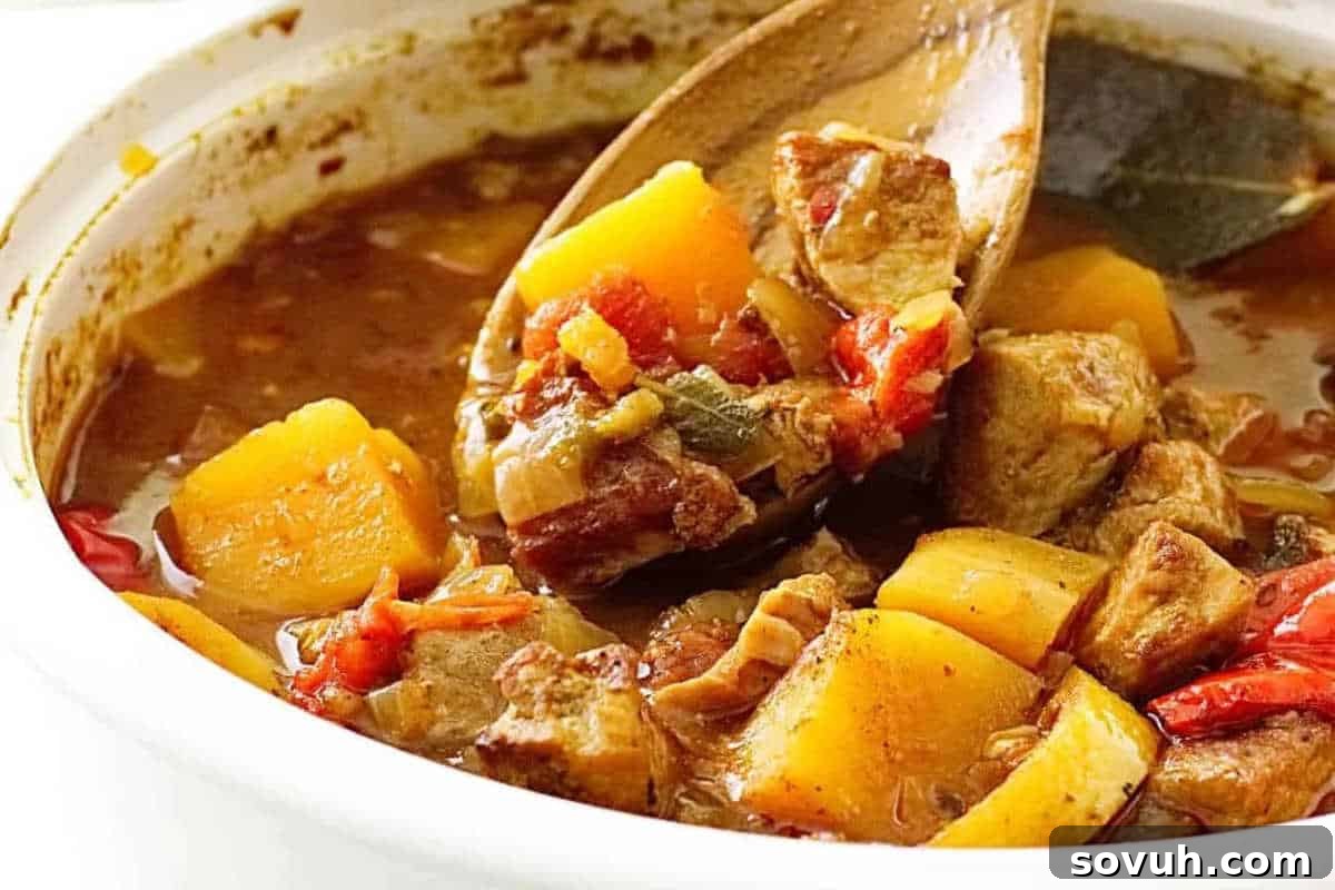 A close-up of a stew with chunks of meat, potatoes, and tomatoes in a brown broth, being stirred with a wooden spoon.