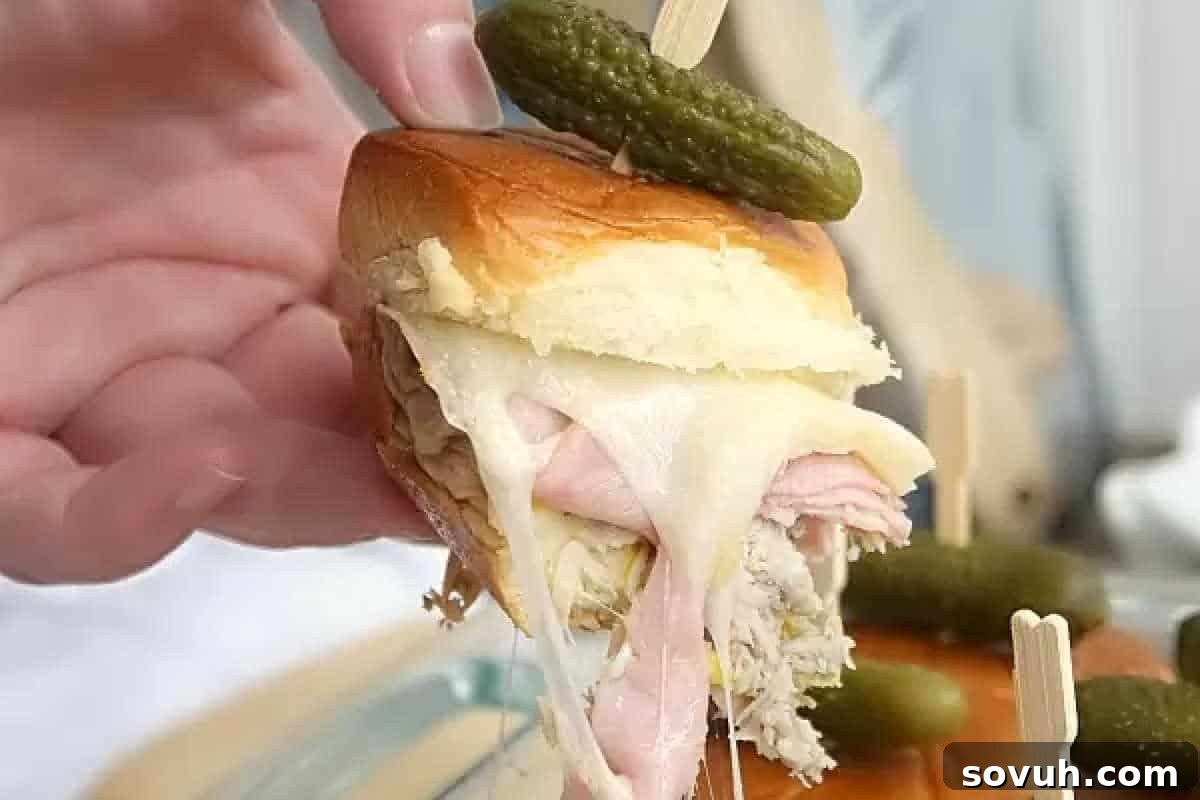 A close-up of a mini sandwich topped with a pickle on a skewer, filled with various meats and melted cheese, being held by a hand.