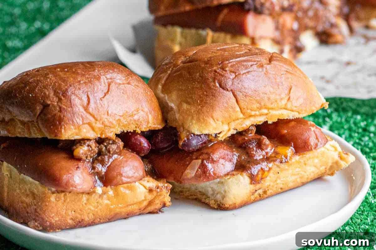 Two chili dogs in soft buns on a white plate. Ground beef and kidney beans are visible in the chili topping. Another dish is visible in the background.