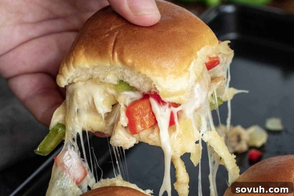 A hand holds a bitten cheesy slider sandwich filled with bell peppers and onions, showing melted cheese stretching from the bite.
