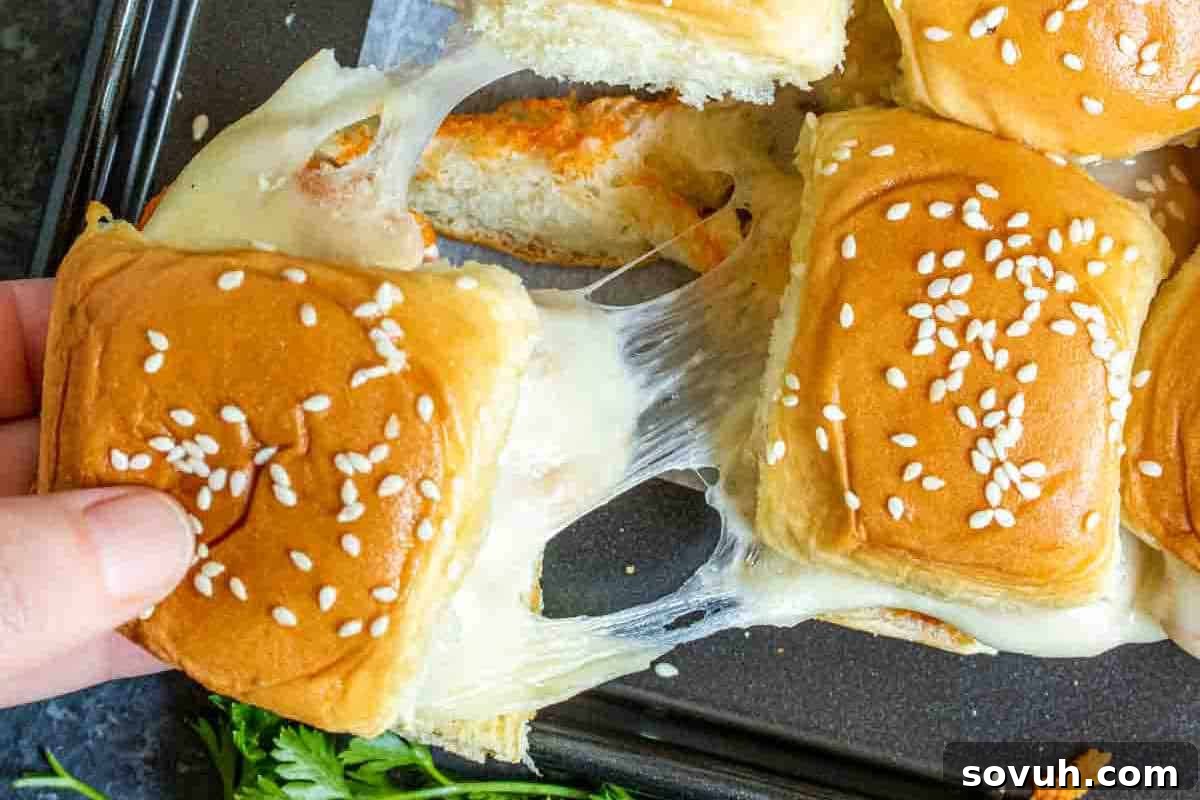 A hand pulling apart a cheesy slider with a golden bun topped with sesame seeds, alongside other similar sliders on a baking tray.