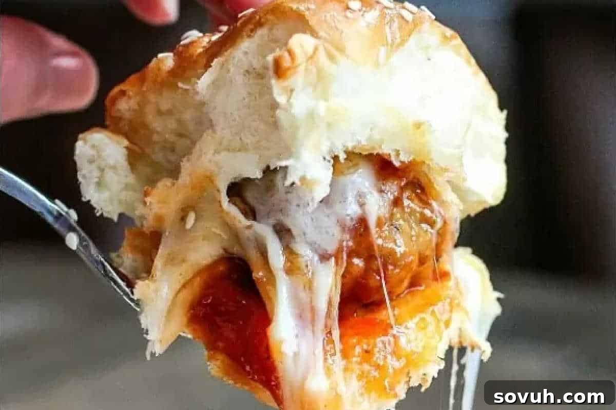 A hand holds a meatball slider sandwich with melted cheese and marinara sauce oozing out, highlighting its cheesy and savory contents.