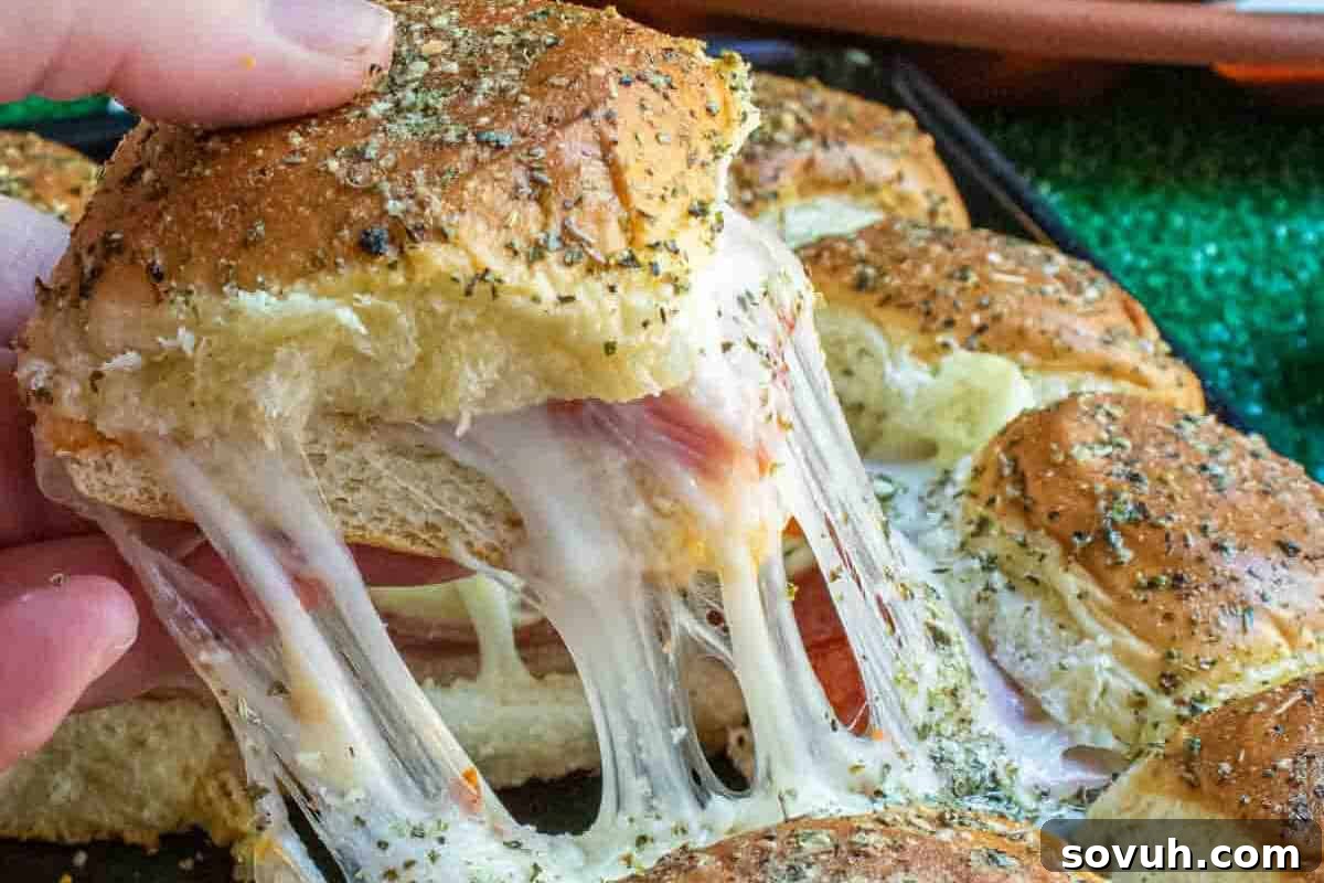 A hand pulling apart a cheesy garlic bread slider. The sandwich filling is visibly stretching, with melted cheese and layers of ham. The bread is topped with herbs.