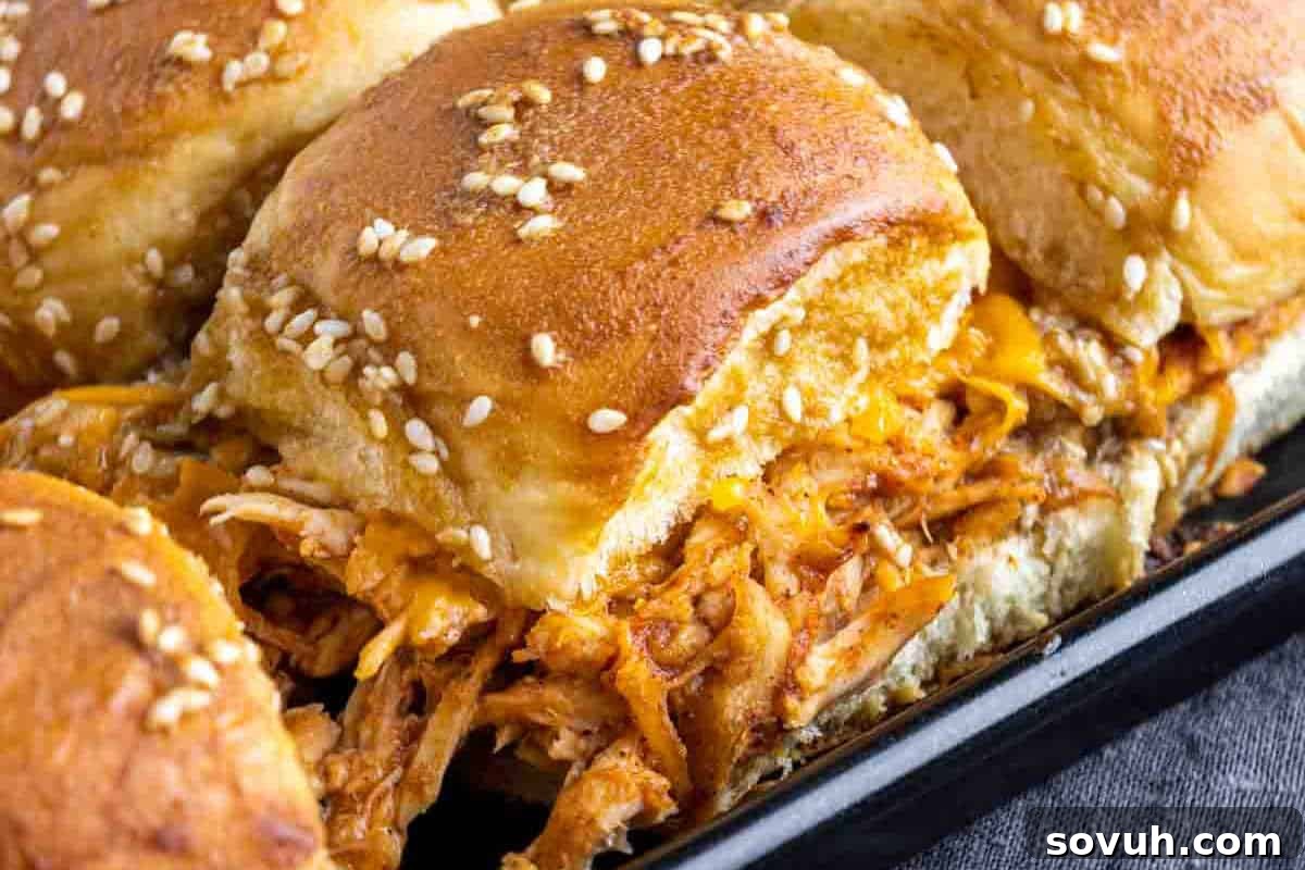 Close-up of a tray of pulled chicken sliders with melted cheese and sesame seed-topped buns.