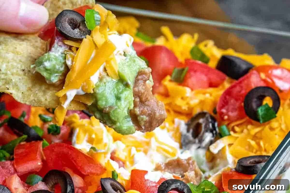 Close-up of a layered dip with shredded cheddar, black olives, tomatoes, green onions, guacamole, and refried beans on a chip.