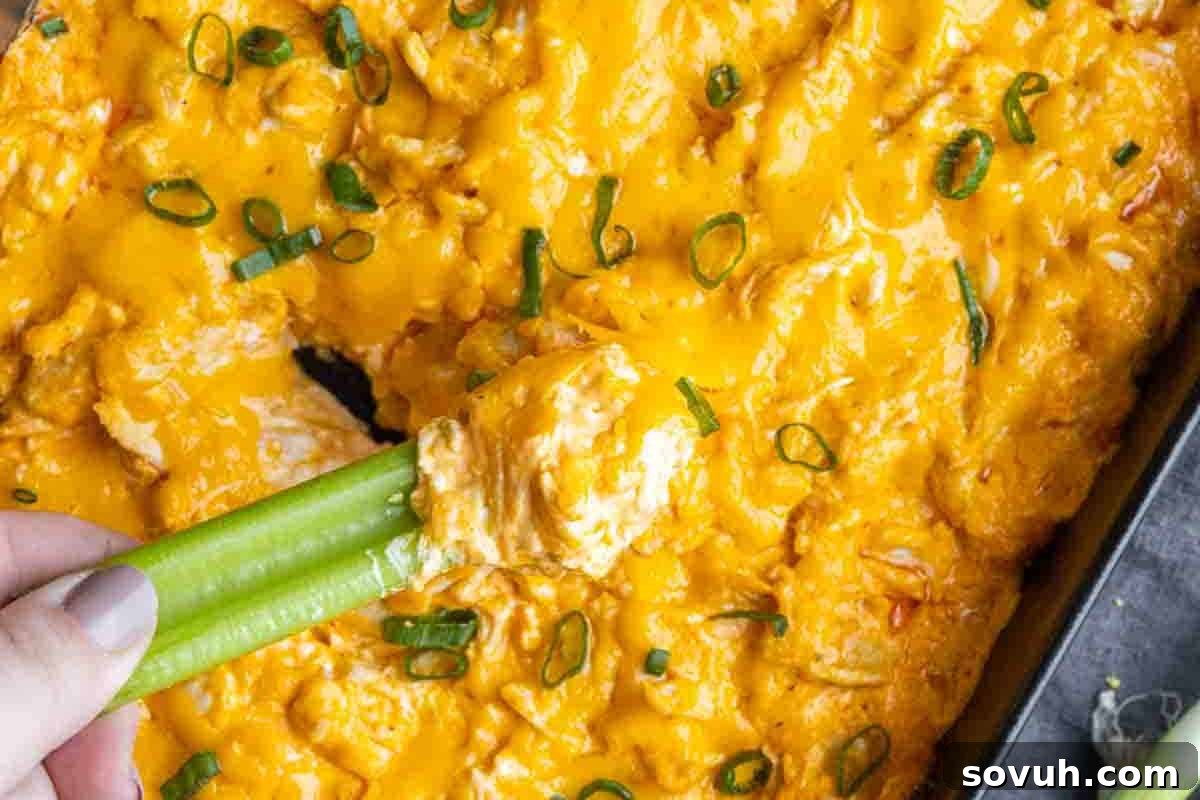 Close-up of a cheesy buffalo chicken dip topped with chopped green onions, with a hand holding celery sticks dipped into it.