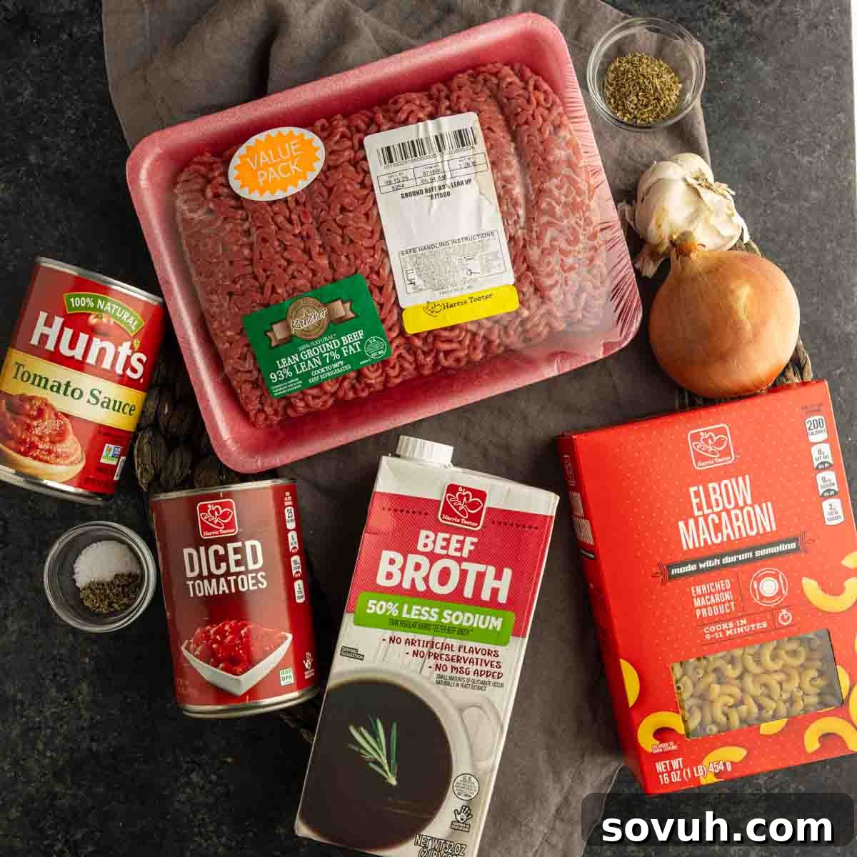 Ingredients for a classic ground beef pasta recipe, perfect for old fashioned goulash, featuring elbow macaroni, beef broth, diced tomatoes, tomato sauce, onion, garlic, and dried herbs arranged on a dark surface.
