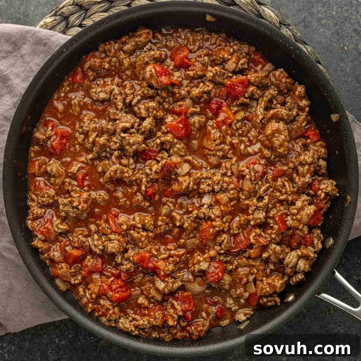 Ground beef cooked with diced tomatoes and sauce in a black skillet, resting on a gray towel and woven placemat—classic American goulash at its best.