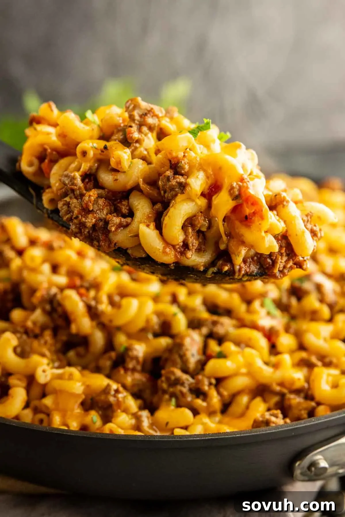 A close-up of a spoonful of cheesy macaroni with ground beef being lifted from a skillet filled with American goulash, showcasing the comforting classic pasta dish.