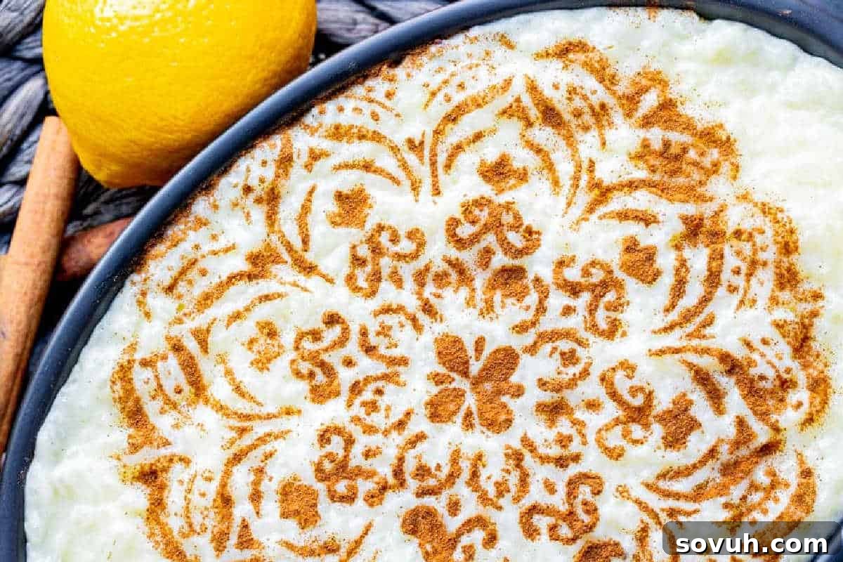 A bowl of creamy Portuguese Rice Pudding (Arroz Doce) adorned with an intricate cinnamon pattern, alongside fresh lemon and a cinnamon stick.