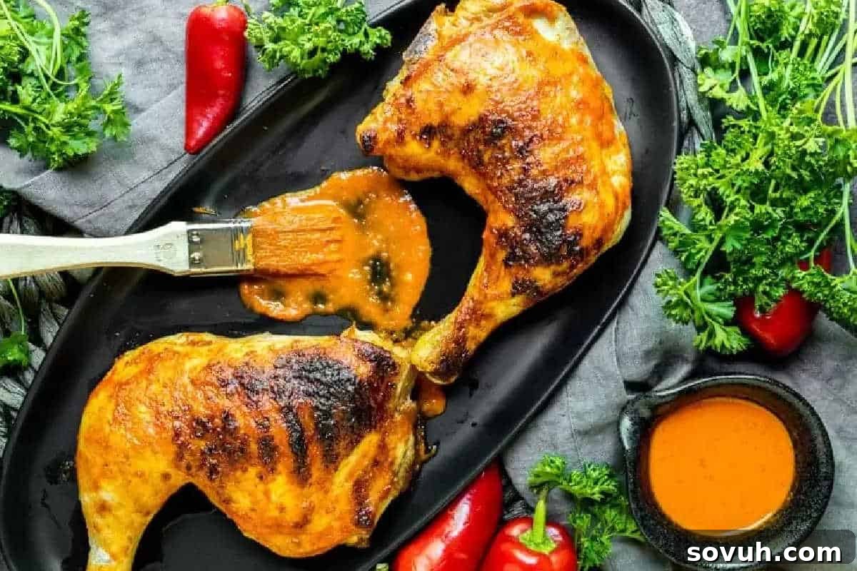 Two grilled Piri Piri chicken leg quarters, brushed with a vibrant orange sauce, surrounded by fresh herbs and chili peppers on a black plate.