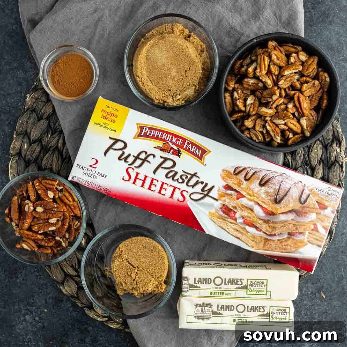 Ingredients for easy sticky buns: puff pastry, brown sugar, pecans, butter, and cinnamon on a textured mat.