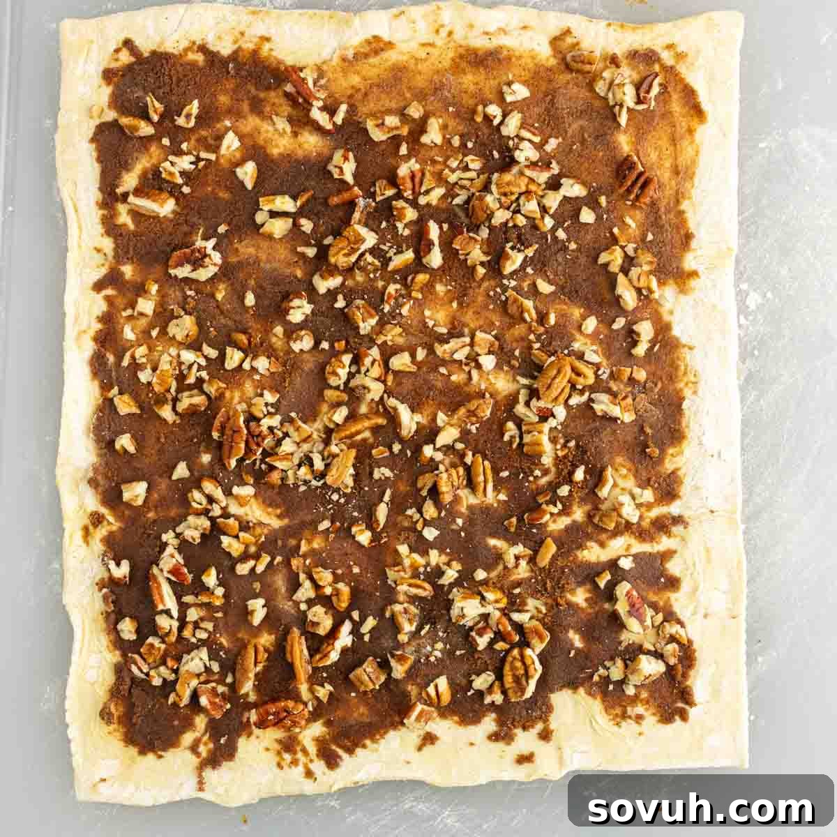 A sheet of puff pastry dough topped with brown sugar, cinnamon, and chopped pecans, ready to be rolled for easy sticky buns.