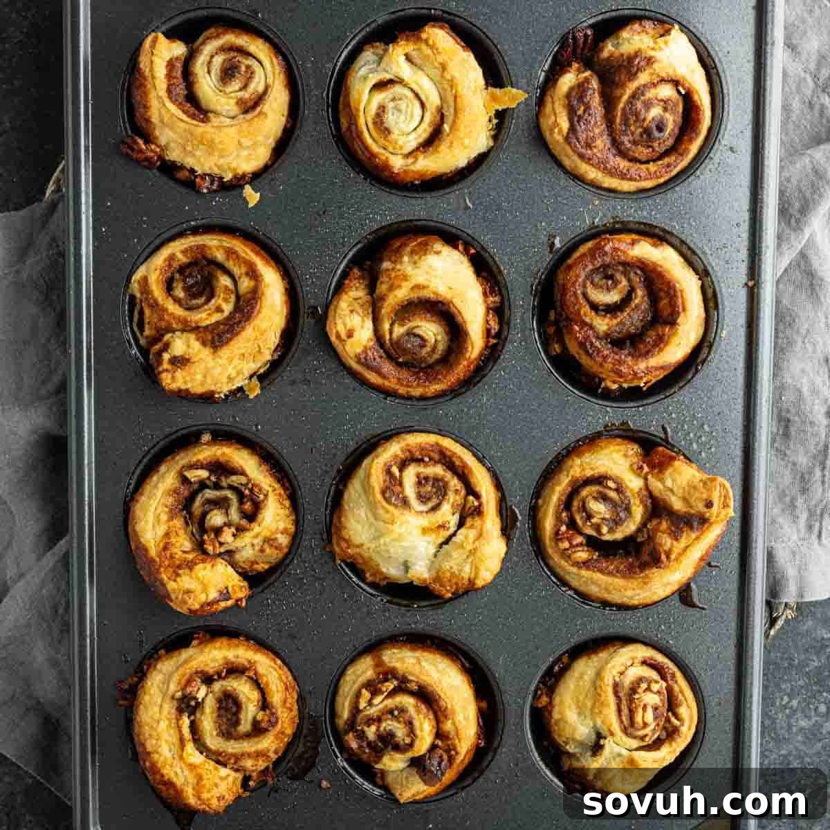 A muffin tray filled with twelve baked puff pastry sticky buns, golden brown and puffed, viewed from above.