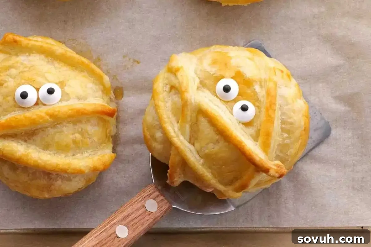 Two baked pastry mummies on parchment paper, each with googly eyes and puff pastry strips as wrappings, one on a spatula.