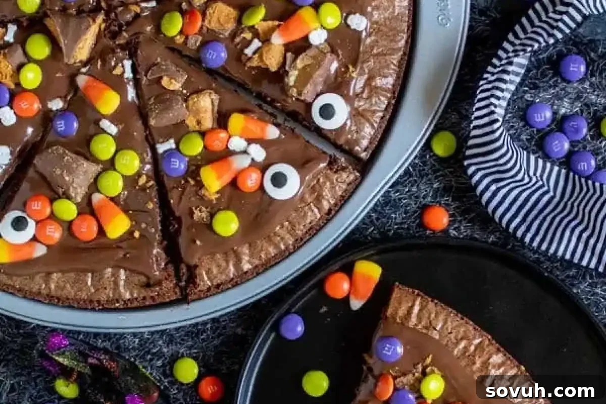 Chocolate pizza topped with candy corn, colorful candies, and edible eyes on a gray surface with a slice on a separate plate. Striped cloth nearby.