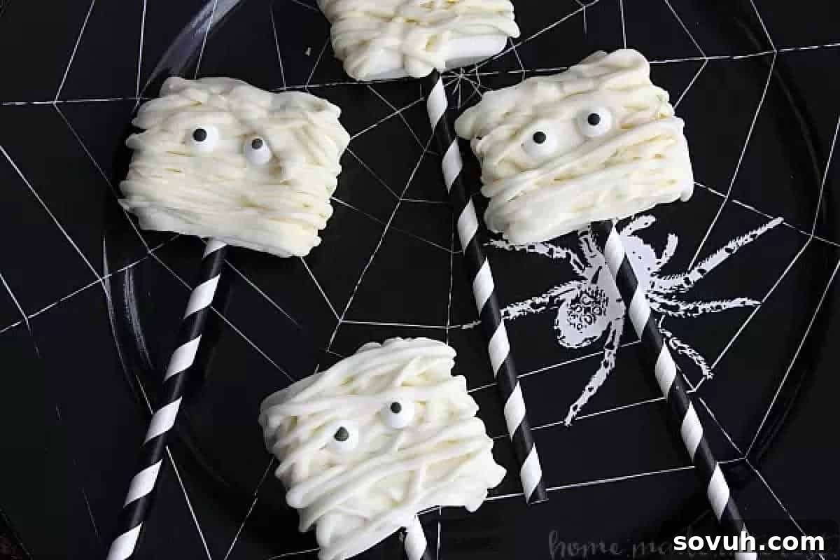 Four marshmallow pops decorated as mummies with white chocolate and candy eyes on a spider web plate.
