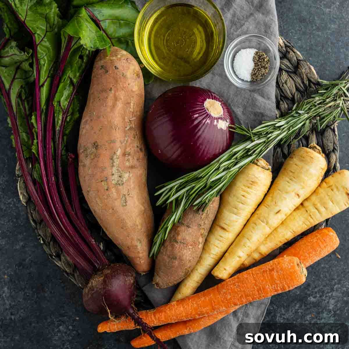 Assorted raw vegetables including sweet potatoes, carrots, parsnips, beet, red onion, and fresh rosemary are prepped for an easy roasted vegetable recipe with oil and seasoning on a woven mat.