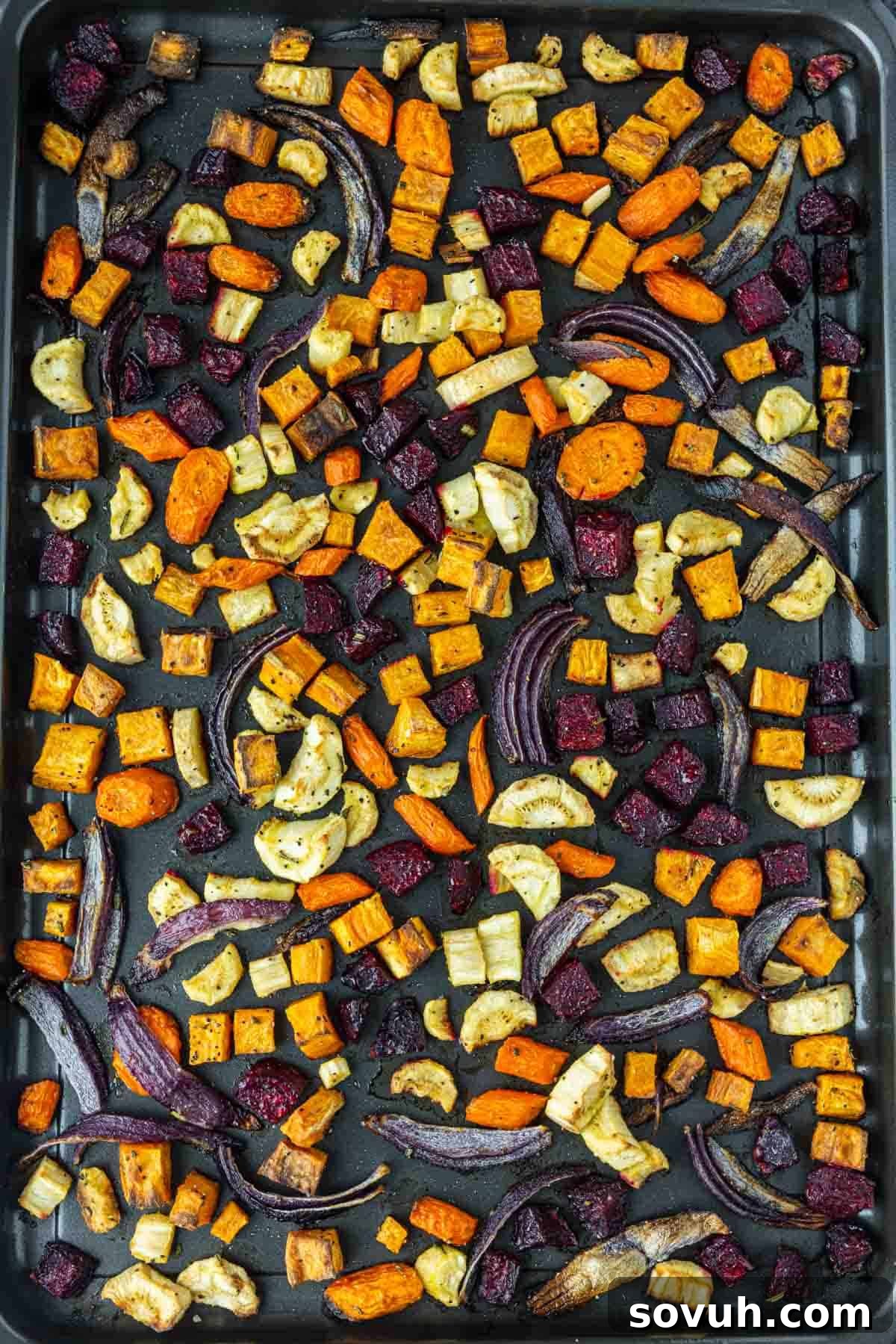 A baking tray filled with an assortment of oven roasted vegetables, including carrots, sweet potatoes, beets, parsnips, and red onion slices, spread out evenly.