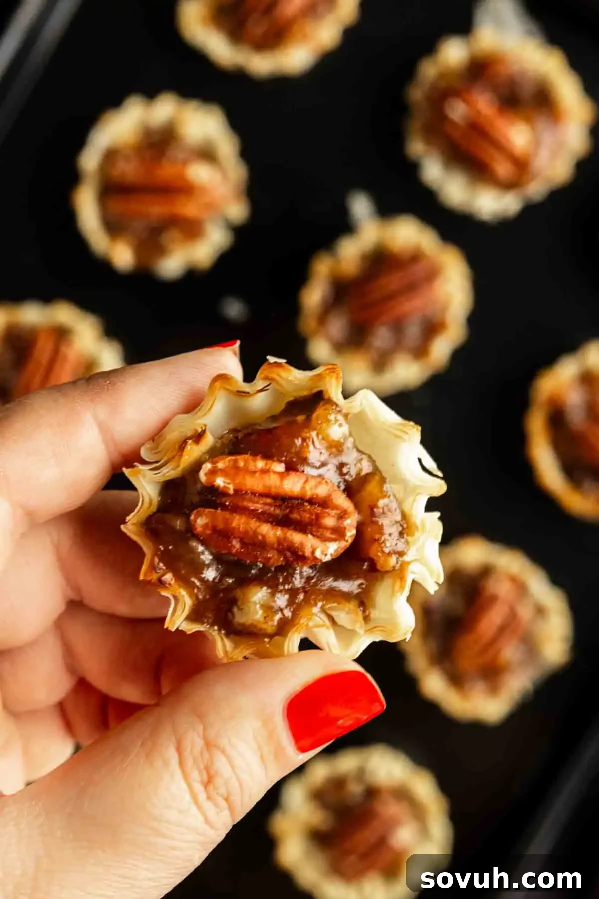 A hand with red nail polish holds a mini pecan pie, featuring a flaky crust and pecan topping; more bite‑sized pecan pie treats are visible on a dark surface in the background.