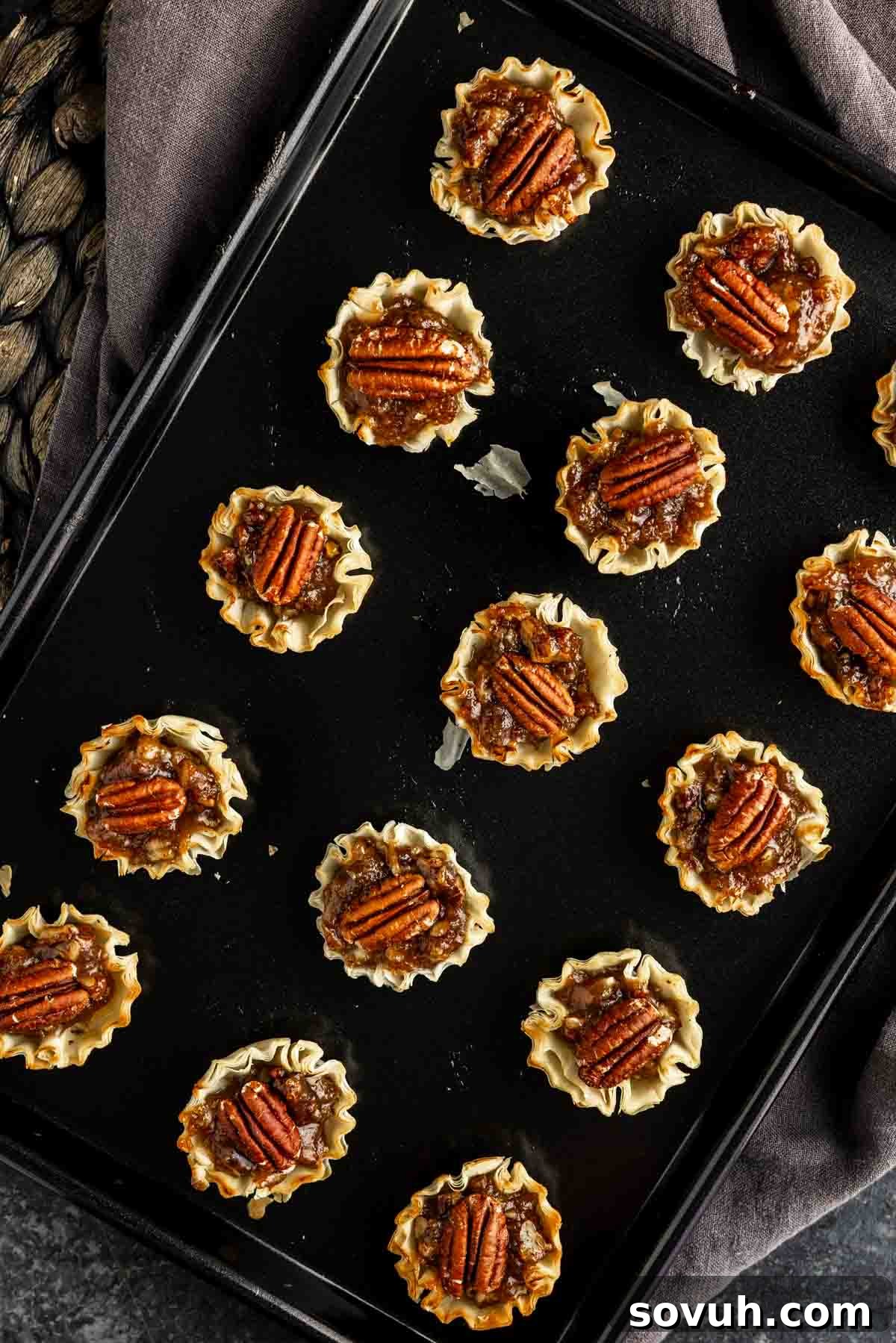 A baking tray filled with bite‑sized pecan pie treats in phyllo dough shells, each topped with pecan halves, viewed from above.