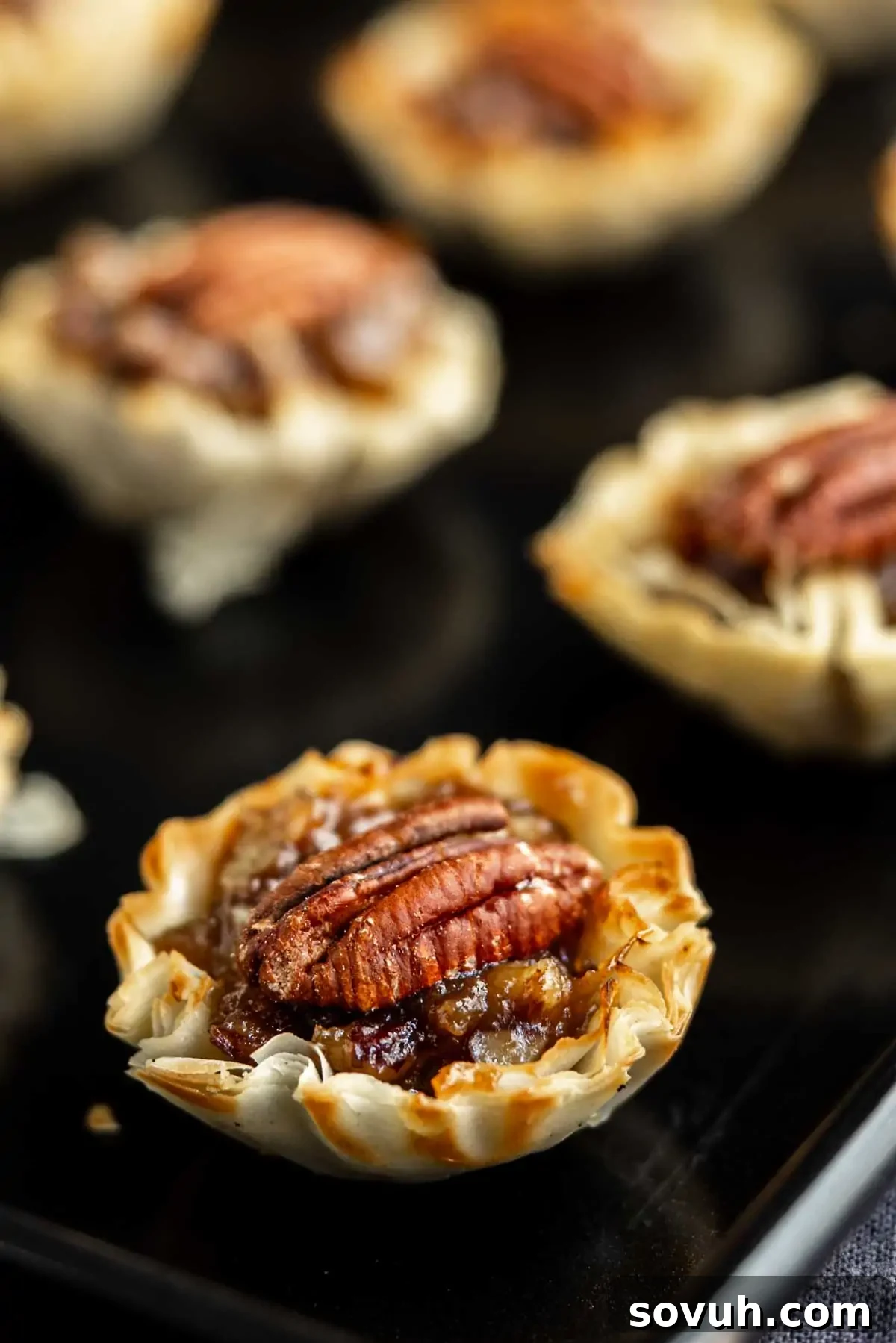 Mini pecan pies with whole pecans on top, baked in flaky phyllo dough cups, make perfect bite-sized pecan pie treats, all arranged on a dark surface.