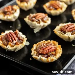 Mini pecan tarts with phyllo pastry shells on a black baking sheet, these bite-sized pecan pie treats are topped with whole pecans and caramelized filling.