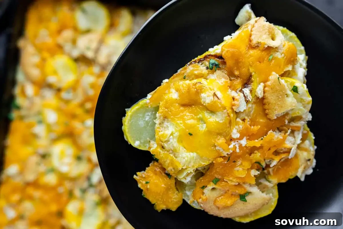 A serving of cheesy yellow squash casserole with a cracker topping on a black plate, next to a baking dish with more casserole.