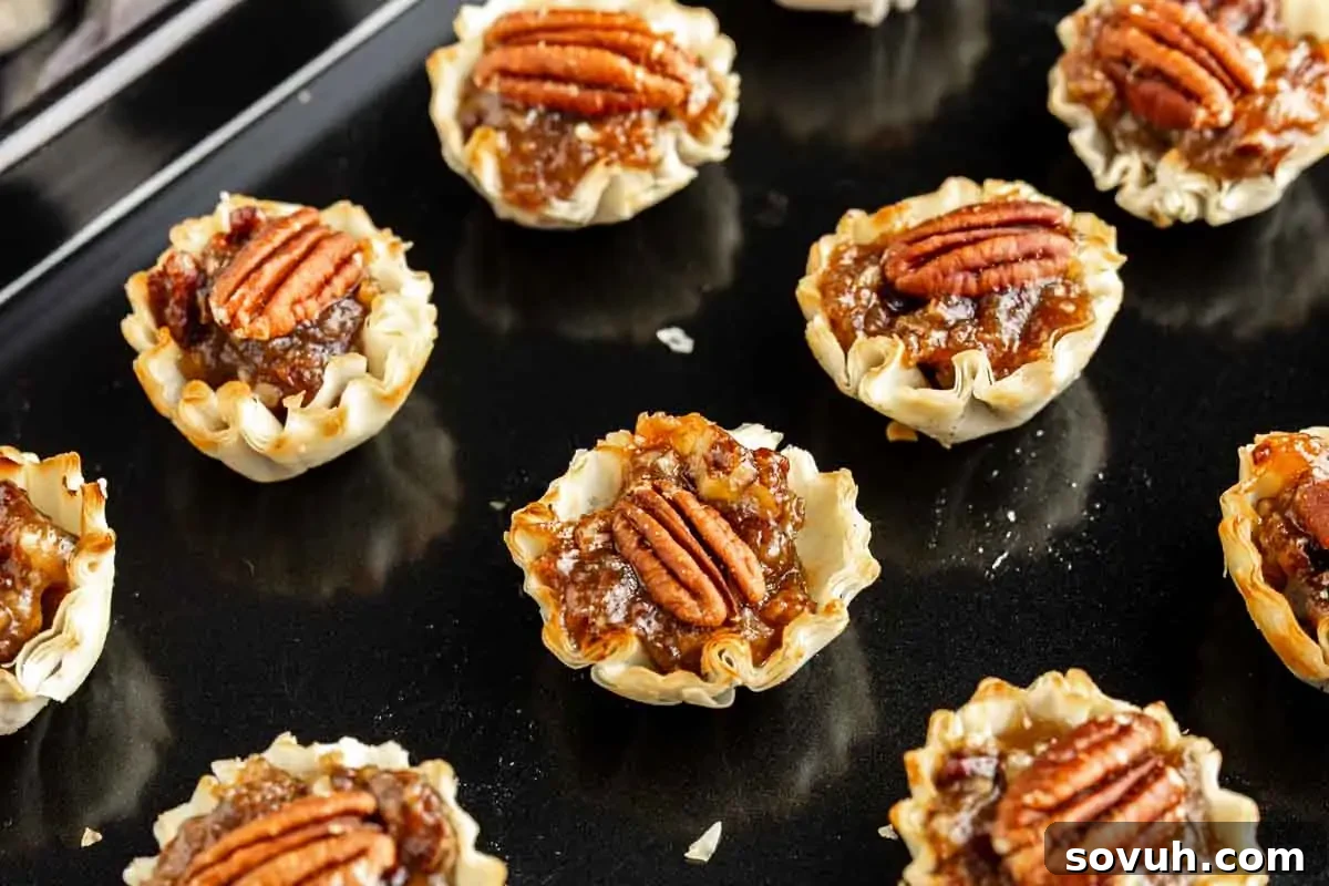 Mini pecan tarts with flaky crusts, filled with pecan pie filling and topped with pecan halves, arranged on a dark baking sheet.