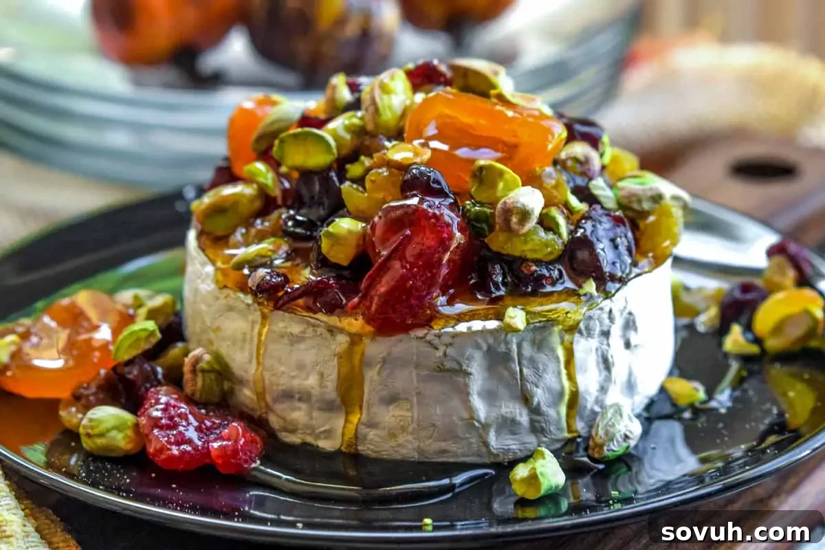 A round of brie cheese topped with dried fruits, pistachios, and honey, served on a black plate with extra toppings around the cheese.