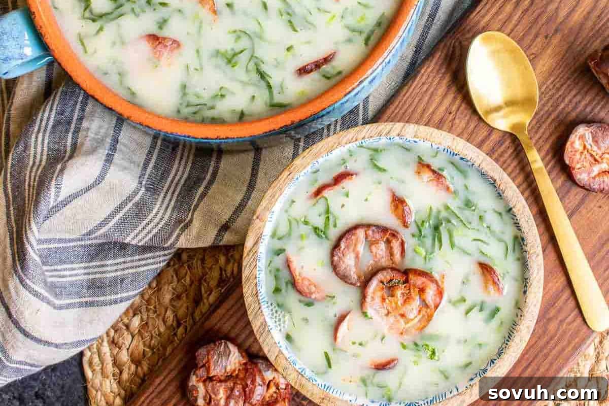 A bowl and pot of creamy soup with slices of sausage and chopped greens, placed on a wooden surface with a gold spoon beside the bowl.