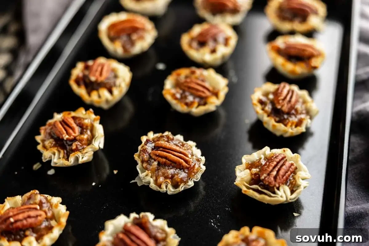 Mini pecan tarts arranged in rows on a black baking sheet, each topped with a whole pecan half and featuring a flaky pastry crust—perfect for easy Thanksgiving desserts and a stress-free Thanksgiving.