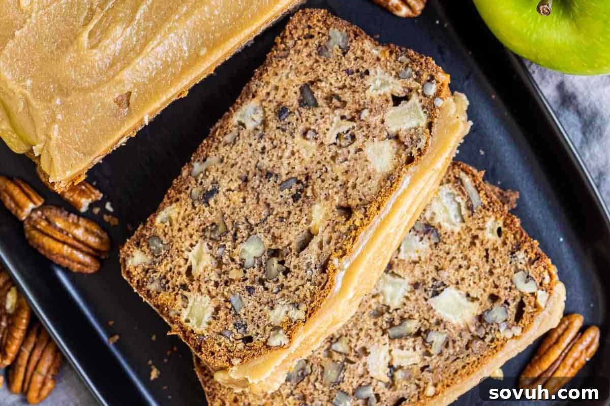 Sliced apple and pecan loaf with caramel frosting on a black tray, surrounded by pecans and a green apple.