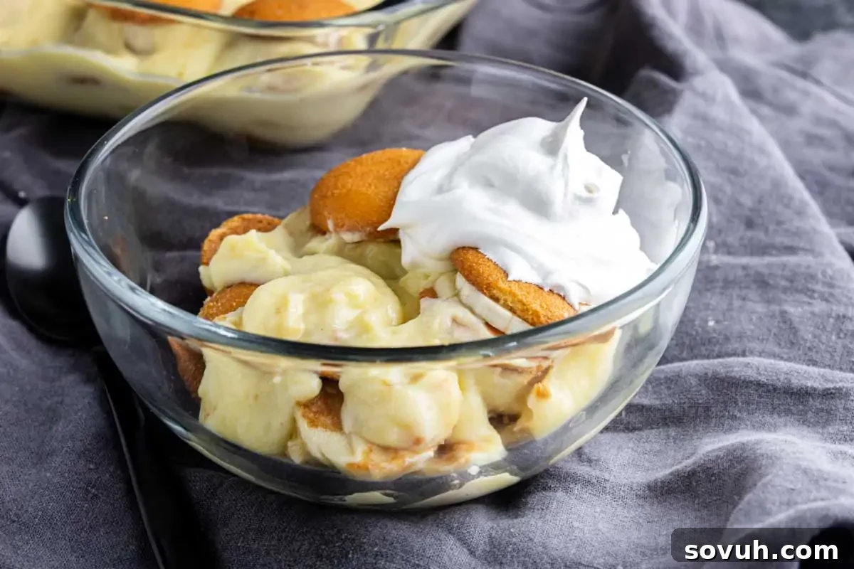 A glass bowl filled with banana pudding, vanilla wafers, and a dollop of whipped cream on top sits on a gray cloth—one of the best easy Thanksgiving desserts for a stress-free holiday treat.