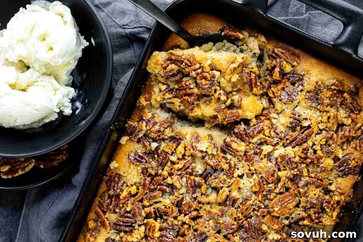 A baked pecan dessert in a rectangular pan with a serving spoon and a bowl of vanilla ice cream beside it.