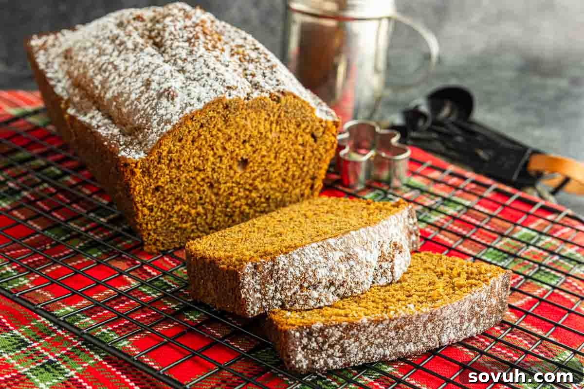A beautifully homemade gingerbread loaf dusted with powdered sugar, with two slices already cut, sits invitingly on a wire rack over a classic red plaid cloth. Subtle baking utensils are visible in the soft-focus background, hinting at the joy of creating this festive treat.