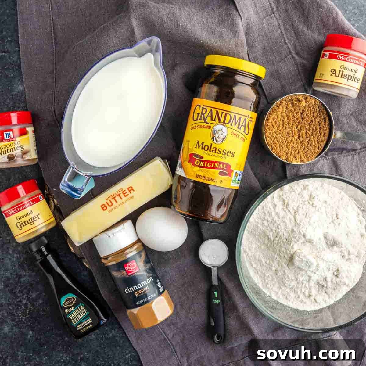 A beautiful flat lay of essential baking ingredients for a gingerbread loaf. Flour, brown sugar, softened butter, fragrant spices, rich molasses, creamy milk, a fresh egg, and pure vanilla extract are arranged on a rustic gray cloth, ready to be transformed into a moist homemade gingerbread cake inspired by the beloved Starbucks recipe.